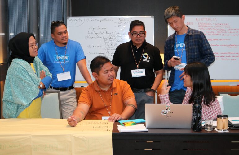 Six people are gathered around a table in a conference room, having a discussion. Some are standing, some are sitting, and there are notes and a laptop on the table with whiteboards full of writing in the background.