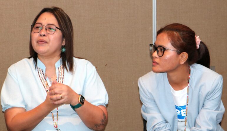 Two women wearing glasses and light blue blazers are sitting indoors in front of a brown wall. One is speaking with hands raised, while the other looks at her attentively.