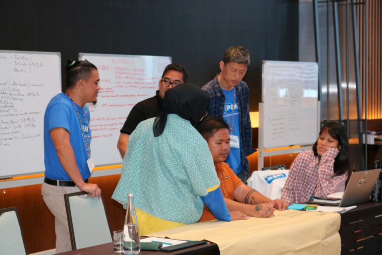 Six people are gathered around a table in a meeting room, engaged in discussion. There are whiteboards with notes and a laptop in the background. Some people are standing while others are seated.
