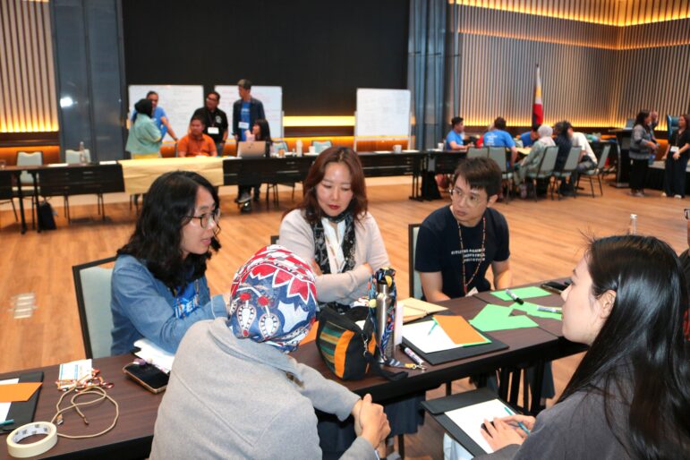 Four people sit at a table engaged in discussion with papers and supplies in front of them. Other groups talk and work in the background in a large, well-lit room with wooden floors and tall, striped walls.