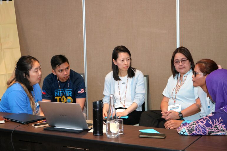 Six people are seated around a table having a discussion. Laptops, water bottles, and papers are on the table. They appear focused and engaged in conversation in a meeting or workshop setting.