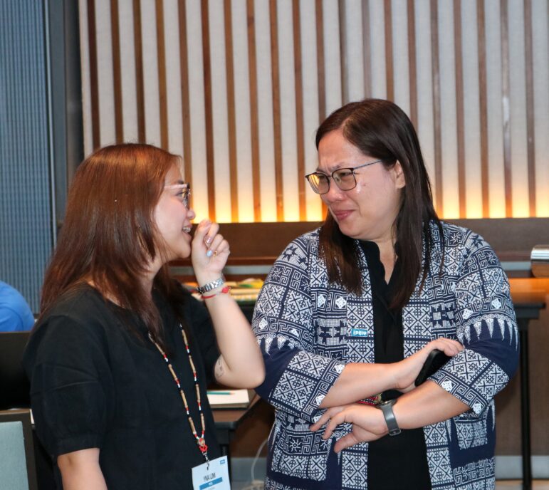 Two women are standing indoors, talking. One woman is smiling and has her hand near her mouth, while the other woman, wearing glasses and a patterned jacket, looks like she is listening closely, with her arms crossed.