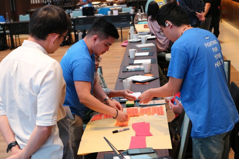 Three people stand around a table covered with paper, sticky notes, and markers in a conference room, collaborating on a project. Other participants and tables with materials are visible in the background.
