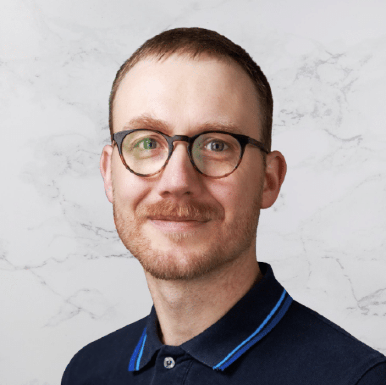 A man with short brown hair, a trimmed beard, and glasses is wearing a navy blue collared shirt with light blue trim. He is facing forward and smiling slightly. The background is a white marble pattern.