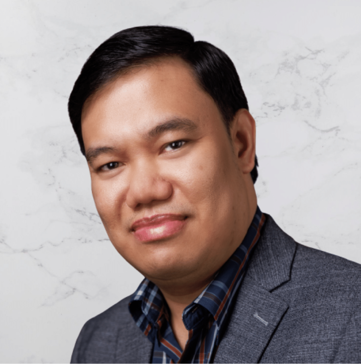 A man with short black hair, wearing a gray blazer over a blue and orange plaid shirt, is looking at the camera with a neutral expression against a light marble background.