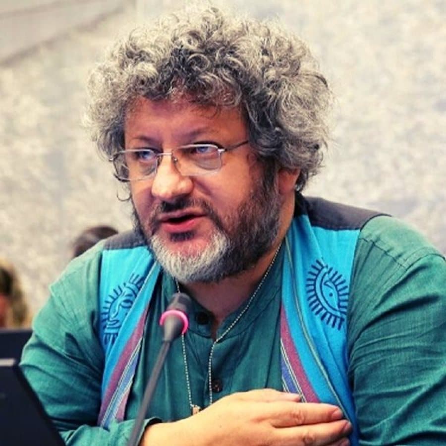 A man with curly gray hair, glasses, and a beard speaks into a microphone. He is wearing a green shirt and a blue vest with decorative patterns, seated at a table with electronic devices in front of him.