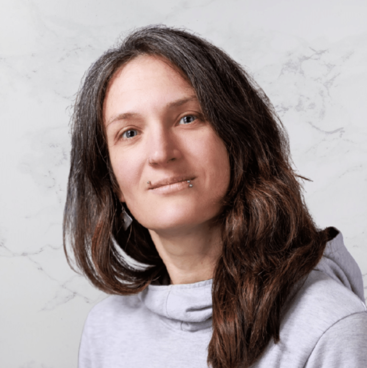 A person with long brown hair with gray streaks, wearing a light gray hoodie, has a neutral expression and a lip piercing. The background is a light, marble-like surface.