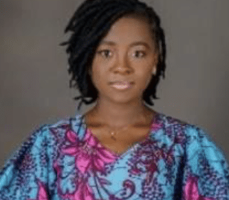 A woman with short, side-swept black hair is wearing a blue and pink patterned top. She is facing forward and looking at the camera, posed against a plain gray background.