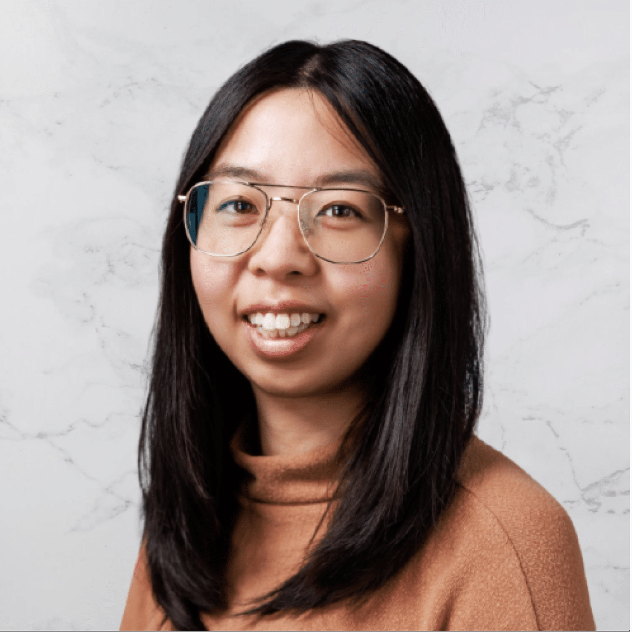 A person with long black hair and glasses smiles at the camera. They are wearing a brown turtleneck sweater and are posed in front of a light marbled background.