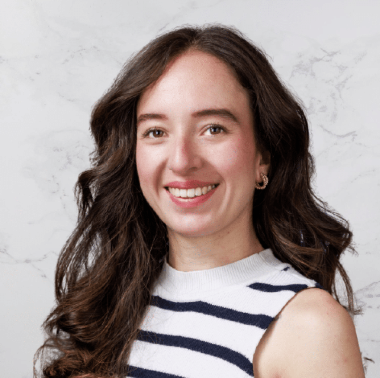 A woman with long, wavy brown hair and fair skin is smiling at the camera. She is wearing a sleeveless white top with navy blue horizontal stripes. The background is a light-colored marble pattern.