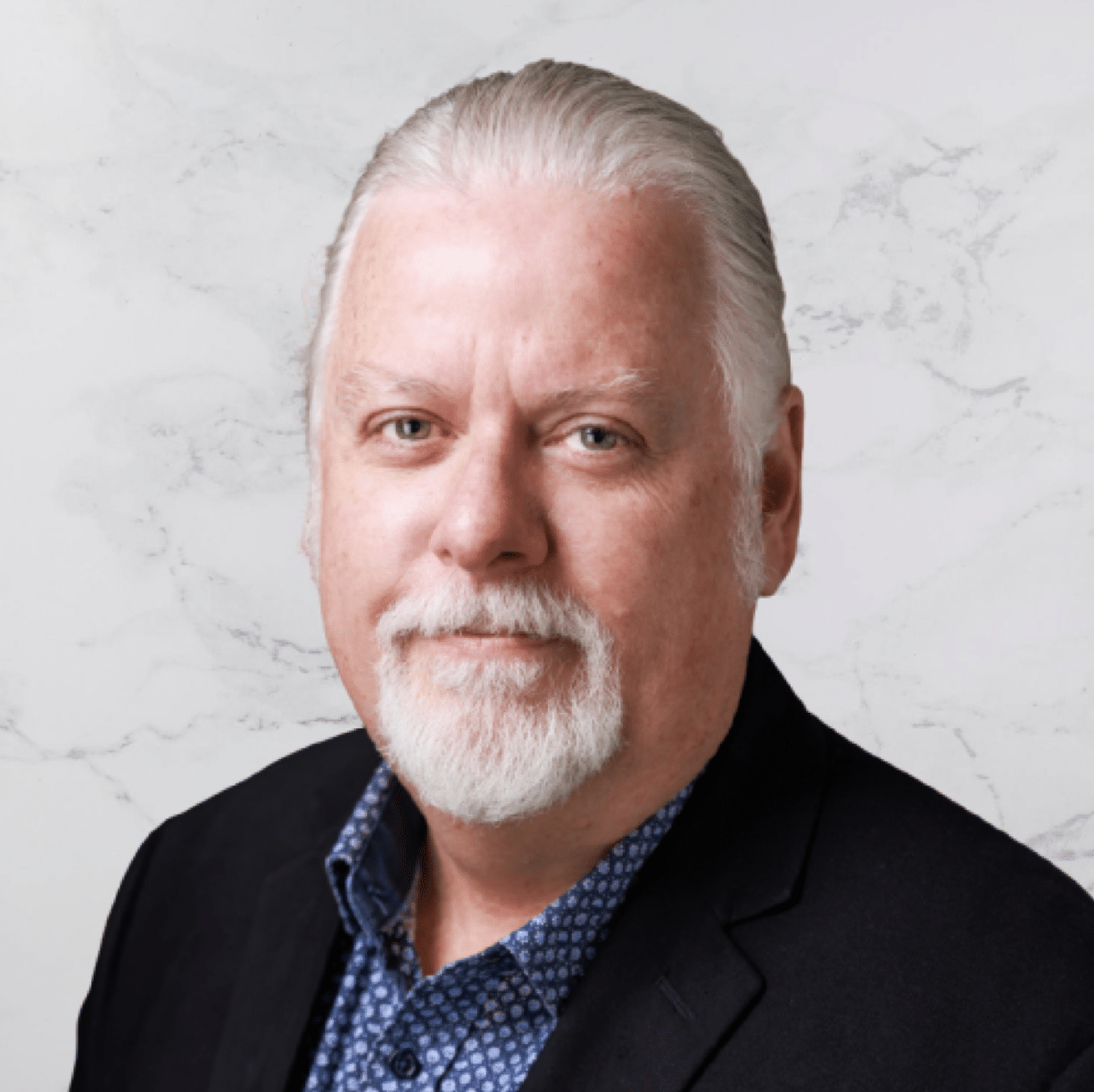 A middle-aged man with white hair and a beard is wearing a dark blazer over a blue patterned shirt. He is facing the camera against a light marble background.