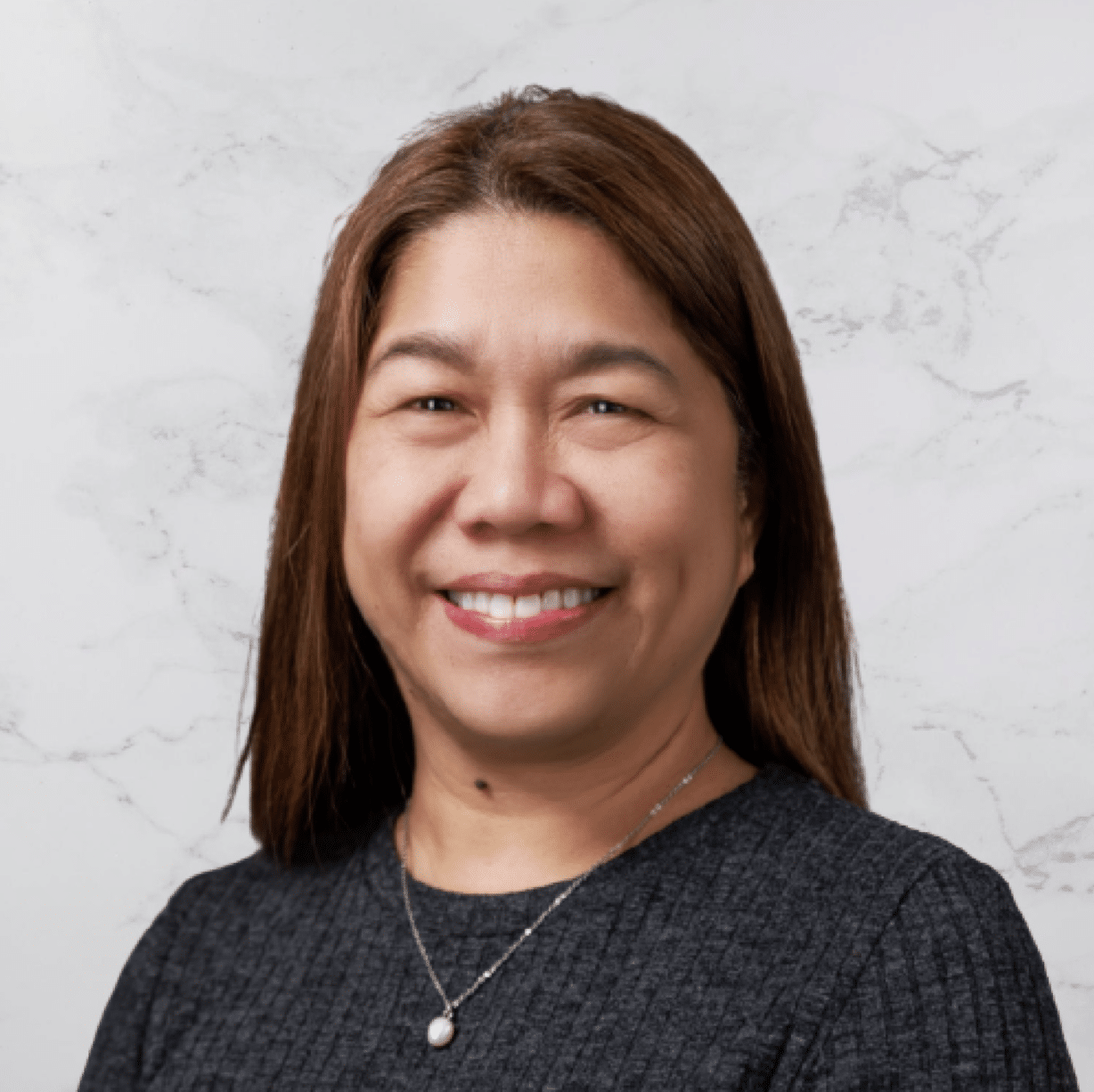 A woman with straight brown hair is smiling at the camera. She is wearing a dark textured top and a necklace with a small pendant. The background is a light-colored marble pattern.