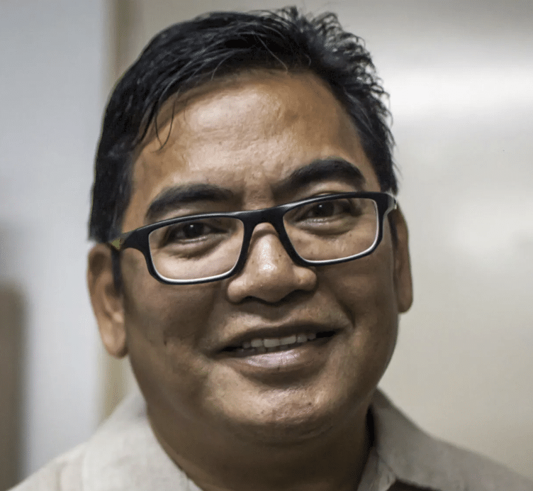 A man with short dark hair and glasses is smiling at the camera. He is wearing a light-colored collared shirt and is standing indoors against a plain background.