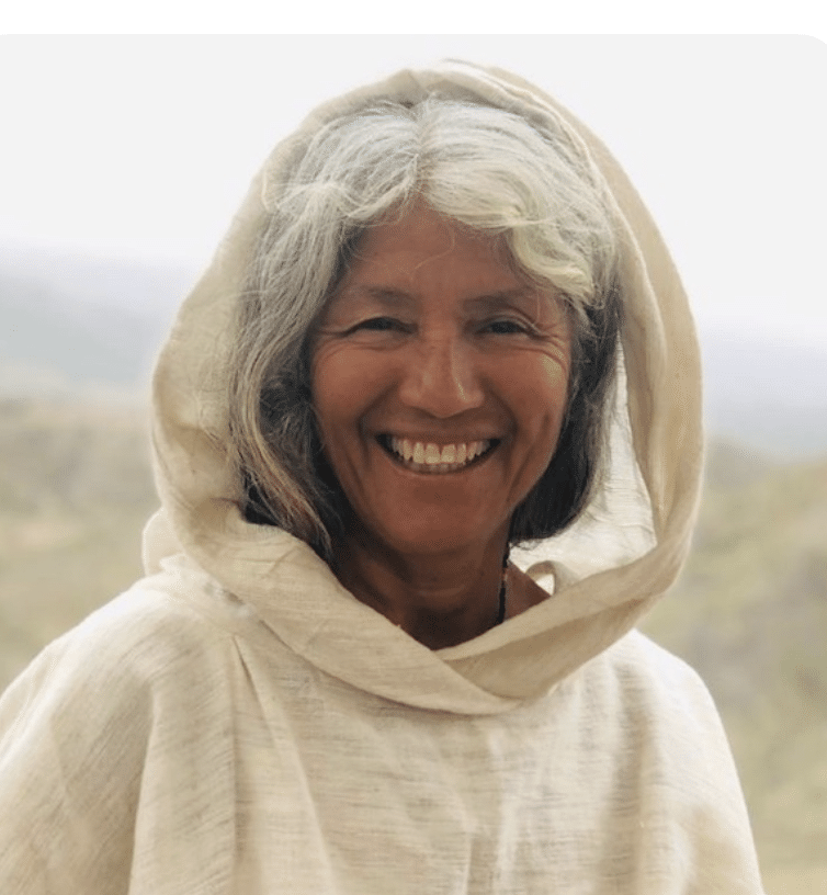 A woman with gray hair and a beige hooded garment stands outdoors, smiling at the camera. The background is blurred, showing a natural, hilly landscape.