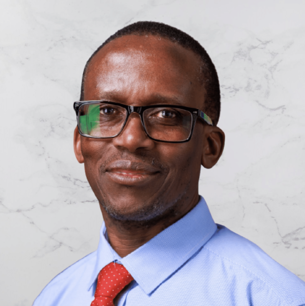 A man wearing glasses, a light blue shirt, and a red tie poses in front of a white marble-patterned background. He is looking slightly to the side and smiling gently.
