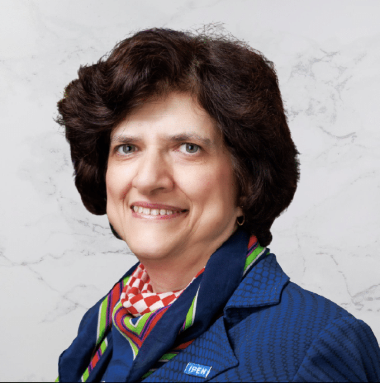 A person with short, dark, curly hair is smiling and wearing a blue blazer, a colorful scarf, and a pin labeled IPEN against a light marble background.