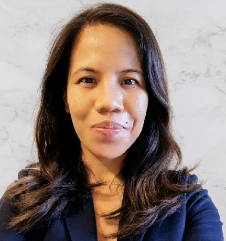 A woman with long dark hair wearing a navy blazer is standing in front of a light-colored, marble-patterned background, looking directly at the camera with a neutral expression.