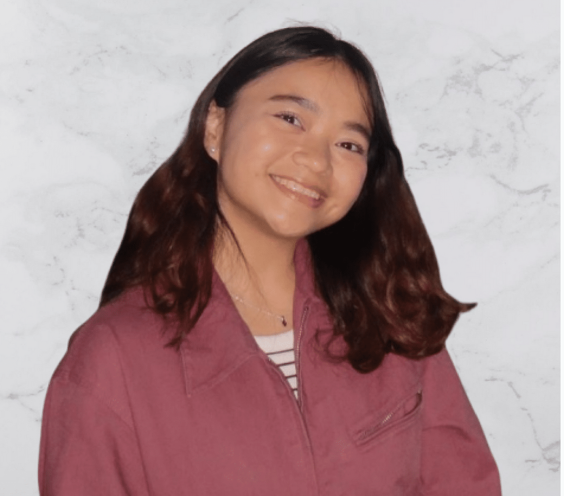 A young person with shoulder-length brown hair smiles at the camera. They are wearing a mauve jacket over a striped shirt, standing in front of a light marble-patterned background.