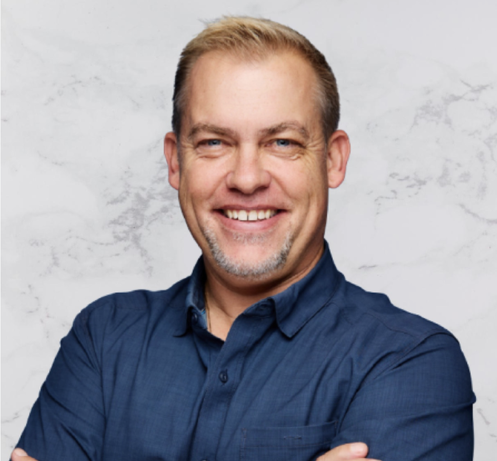 A middle-aged man with short blond hair and a trimmed beard smiles at the camera. He is wearing a blue button-up shirt and stands in front of a light gray, marbled background with his arms crossed.