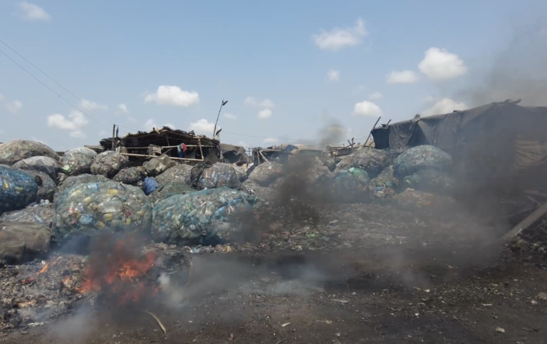 Large piles of plastic waste are stacked outdoors near makeshift structures. Smoke and flames rise from burning debris in the foreground, under a blue sky with scattered clouds.
