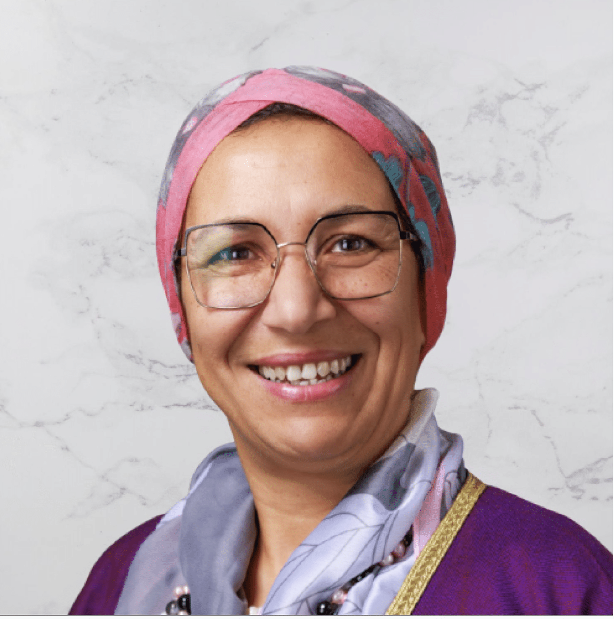 A woman wearing glasses, a pink and gray patterned headscarf, and a purple top smiles at the camera against a light marble-patterned background.