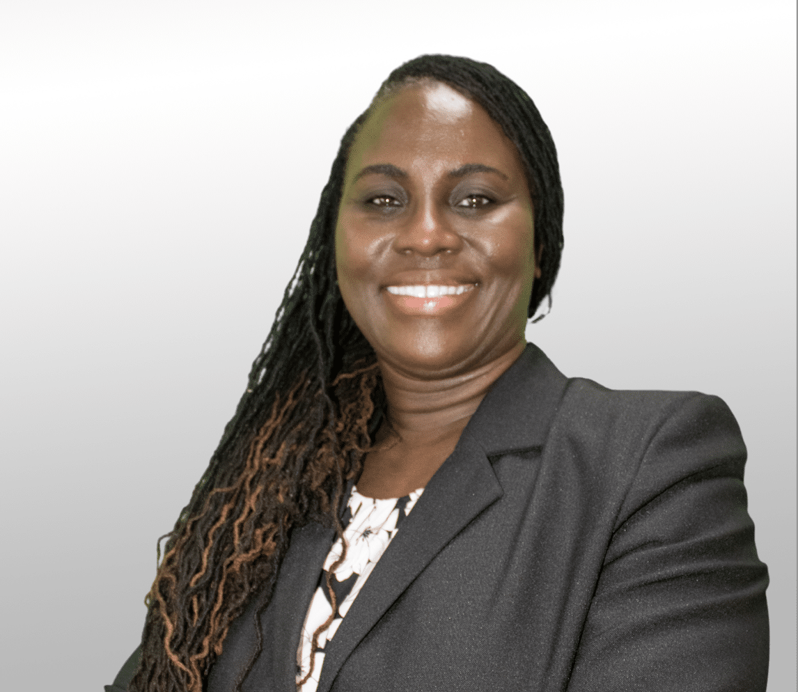 A woman with long, braided hair wearing a dark blazer and patterned top smiles at the camera against a light gray background.