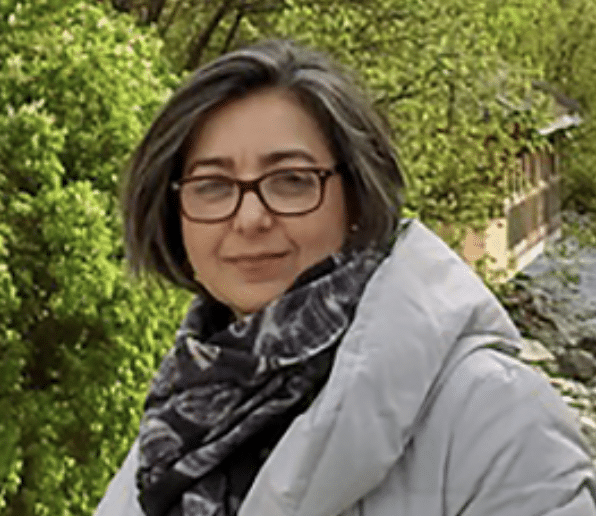 A person with shoulder-length gray hair and glasses is wearing a light-colored coat and a patterned scarf. They are standing outdoors in front of green foliage and a building.