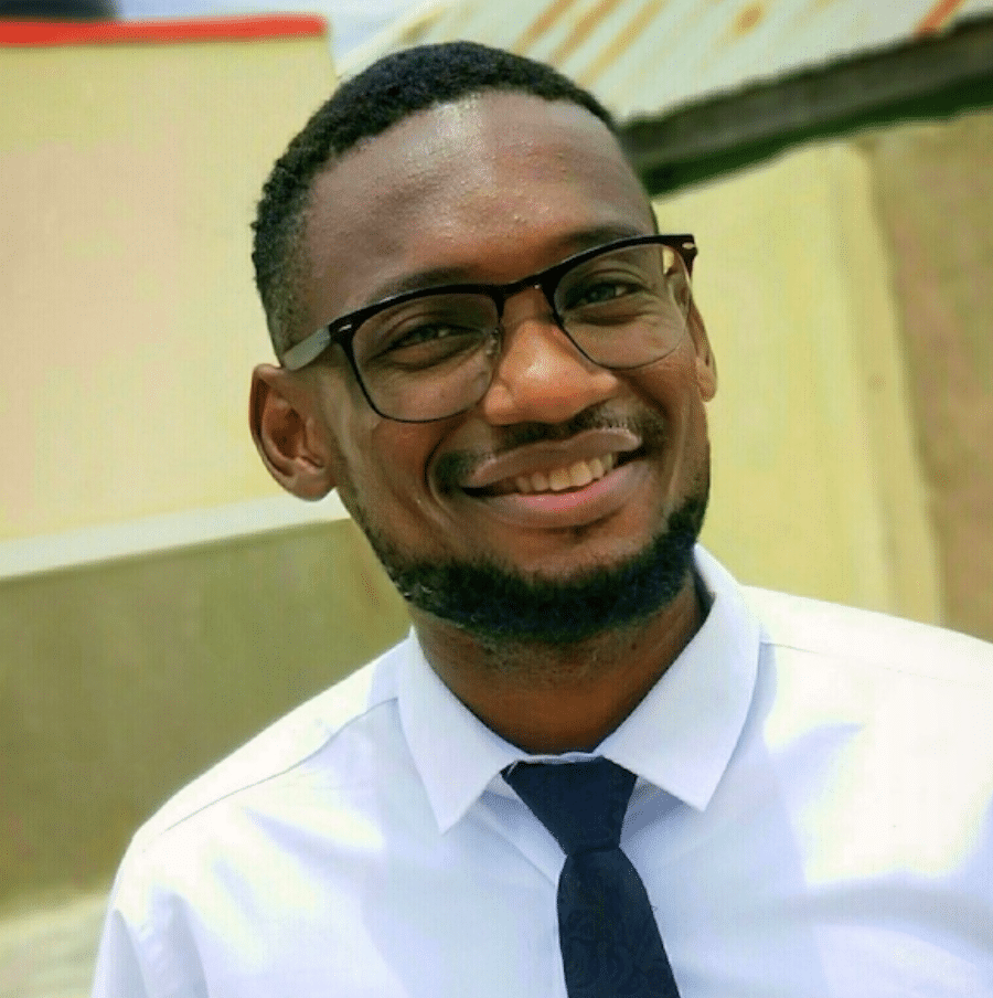 A man with short hair, glasses, and a beard is smiling. He is wearing a white collared shirt and a dark tie, standing outdoors in front of a beige building with a corrugated metal roof.