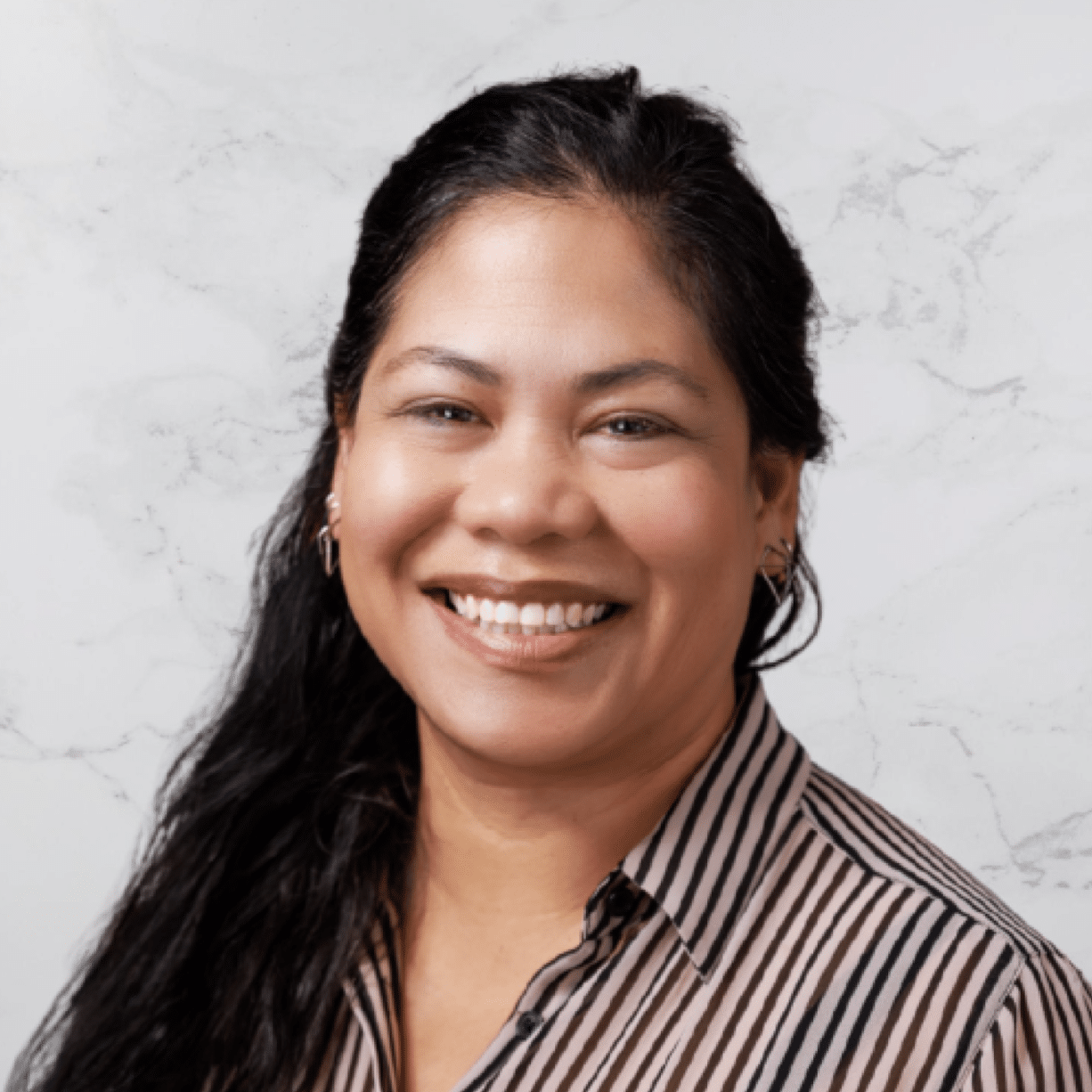 A woman with long, dark hair smiles at the camera. She is wearing a striped button-up shirt and small earrings, posed in front of a light gray, marbled background.