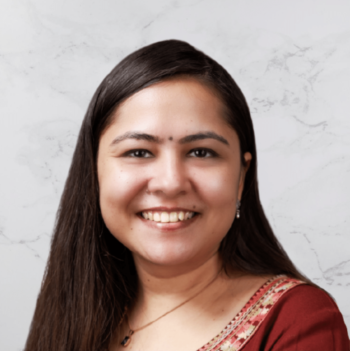 A woman with long dark hair smiles at the camera. She is wearing a traditional outfit with intricate patterns on the neckline. The background is light-colored with a subtle marble texture.