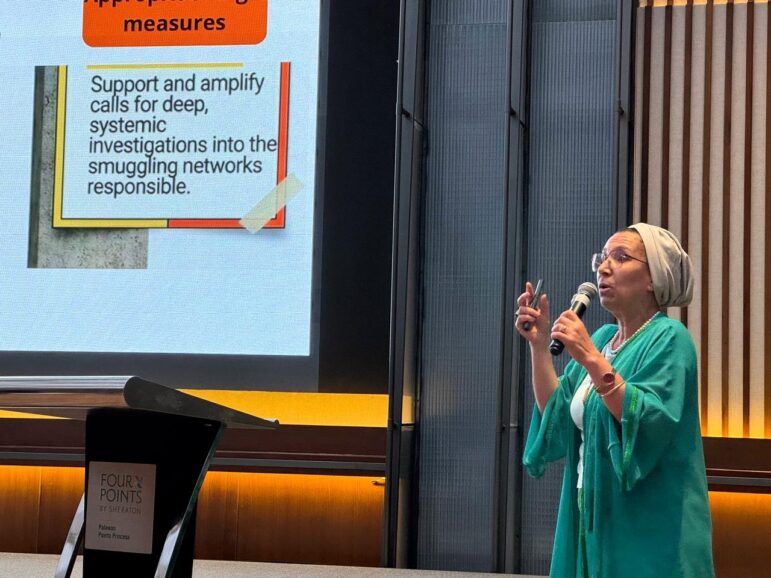 A person in a green outfit and headscarf speaks into a microphone while gesturing with one hand. Behind them is a screen displaying a slide about investigations into smuggling networks. A podium is visible to the left.