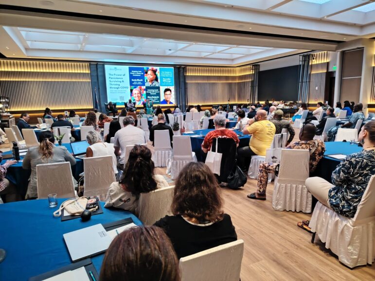 A group of people seated at round tables in a conference room face a stage with a panel discussion. A large screen behind the speakers displays information and images of the panelists.
