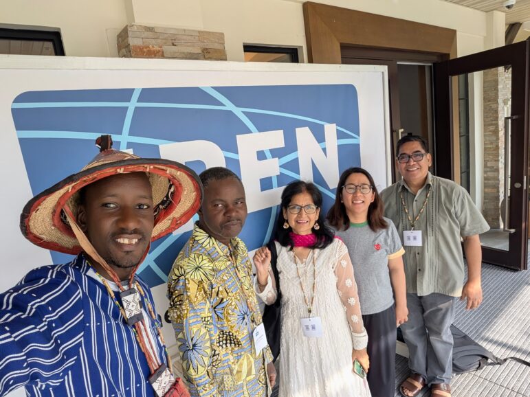 Five people stand and smile in front of a large sign displaying the letters “PEN.” They are dressed in a variety of colorful, cultural, and professional attire. The background features a building entrance.