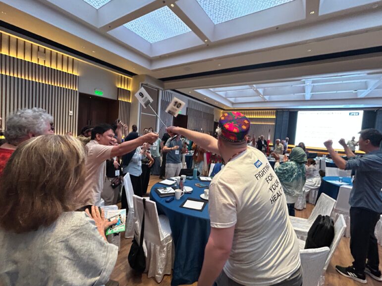 A group of people in a conference room stand around tables, raising signs with numbers. Some are smiling, and one person wears a colorful cap and a T-shirt reading “FIGHT FOR YOUR HEALTH.” The room has high ceilings and bright lighting.