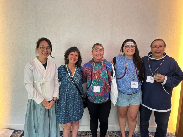 Five people stand side by side, smiling at the camera. They wear a mix of traditional and modern clothing, each with a name badge around their necks. The background is a neutral, textured wall with soft lighting.