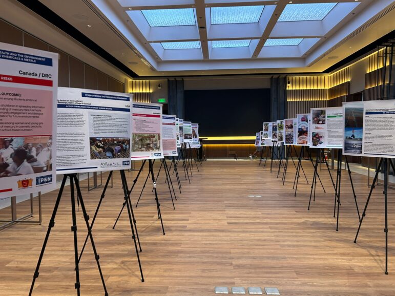 A conference room with large poster boards on tripods arranged in two rows, displaying information and images. The room has a wooden floor, high ceiling with skylights, and recessed lighting along the walls.