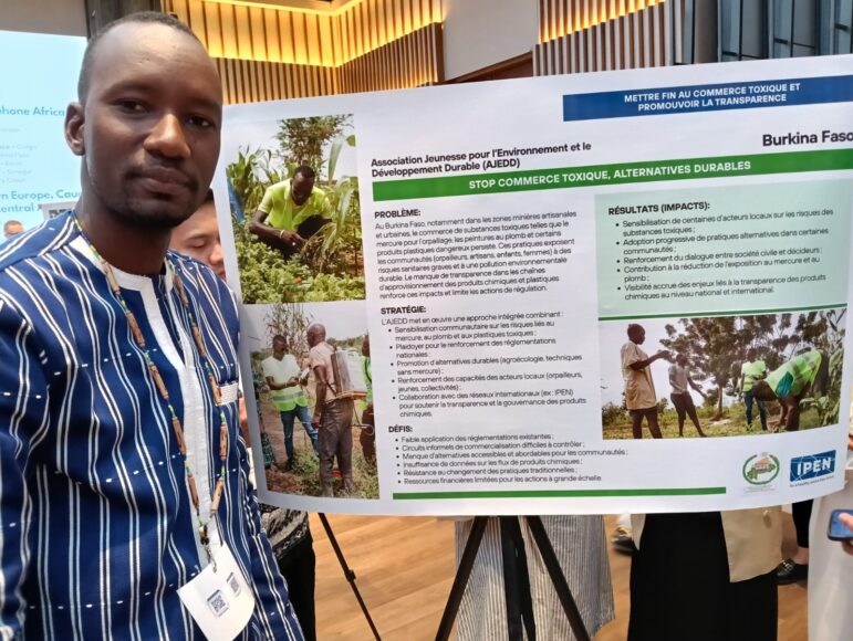 A man in a striped shirt stands beside a display board about sustainable development and stopping toxic trade in Burkina Faso. The board includes text, images of people working in agriculture, and logos from supporting organizations.