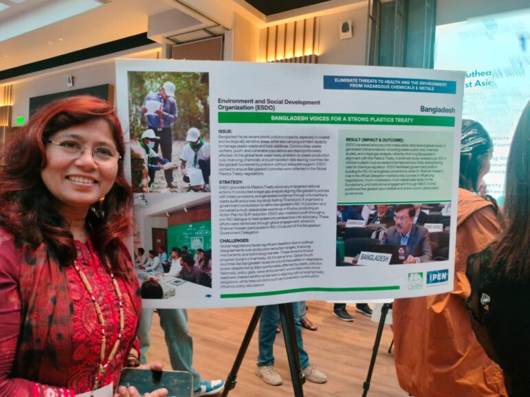 A woman with long reddish hair and glasses stands smiling in front of a poster presentation about Bangladesh’s plastic pollution efforts at an indoor event. The poster contains text, photos, and a headline.