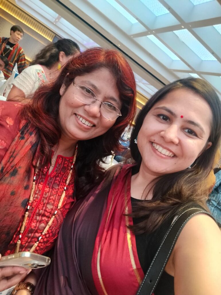 Two women wearing sarees are smiling and posing for a photo at an indoor event. The background shows other people and a brightly lit ceiling.