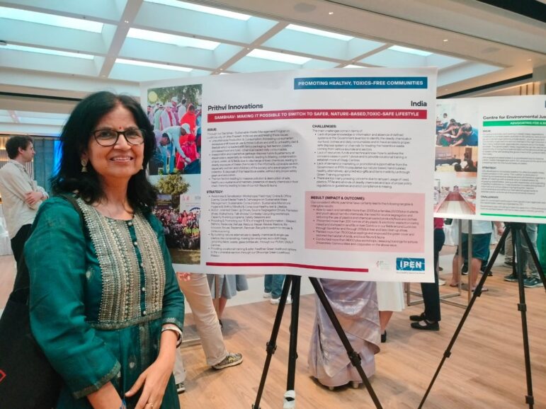 A woman in a green traditional outfit stands smiling next to a display board titled Prithvi Innovations at an indoor event. The board presents information about a project promoting a toxin-free lifestyle in India.