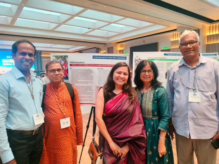 Five people stand indoors in front of research posters at a conference or academic event. They are smiling and dressed in formal or semi-formal attire. The setting is well-lit with a modern ceiling and walls.