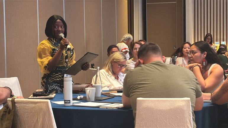 A woman stands and speaks into a microphone while holding a tablet at a round table meeting. Other people are seated around the table, listening and taking notes. The setting appears to be a conference or workshop.