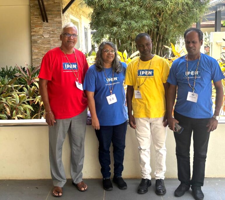 Four adults stand outdoors in front of plants and a building. Two people wear blue IPEN t-shirts, one wears a red IPEN t-shirt, and one wears a yellow IPEN t-shirt. All have name badges on lanyards.