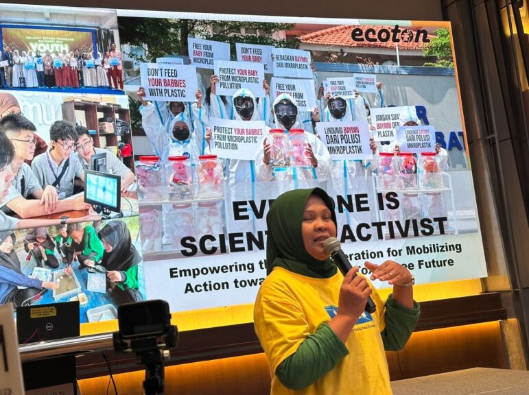 A woman in a yellow shirt speaks into a microphone in front of a large screen displaying a collage of youth holding protest signs about plastic pollution and climate change. The words Everyone is a science activist are visible on the screen.