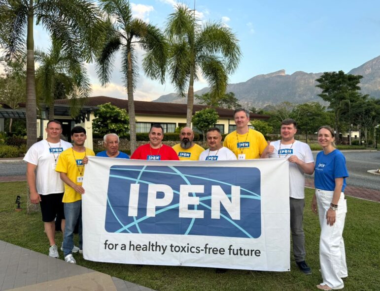 A group of nine people stand outdoors in front of palm trees and mountains, holding a large banner that reads, “IPEN for a healthy toxics-free future.” Most are wearing IPEN shirts in yellow, blue, or white.