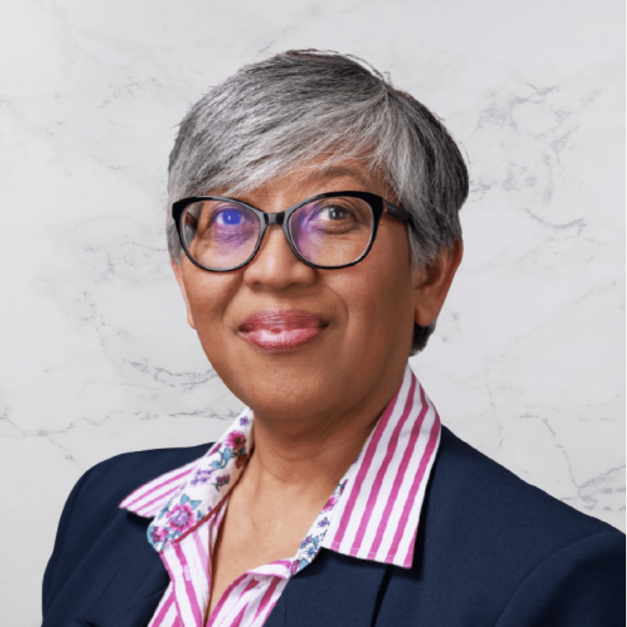 A person with short gray hair and glasses is wearing a navy blazer over a pink and white striped shirt with a floral collar, posing in front of a light marble background.