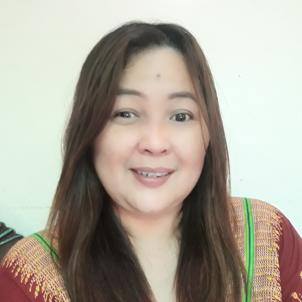 A woman with long brown hair and light skin is smiling at the camera. She is wearing a patterned top with green and yellow accents, and is positioned in front of a plain, light-colored background.