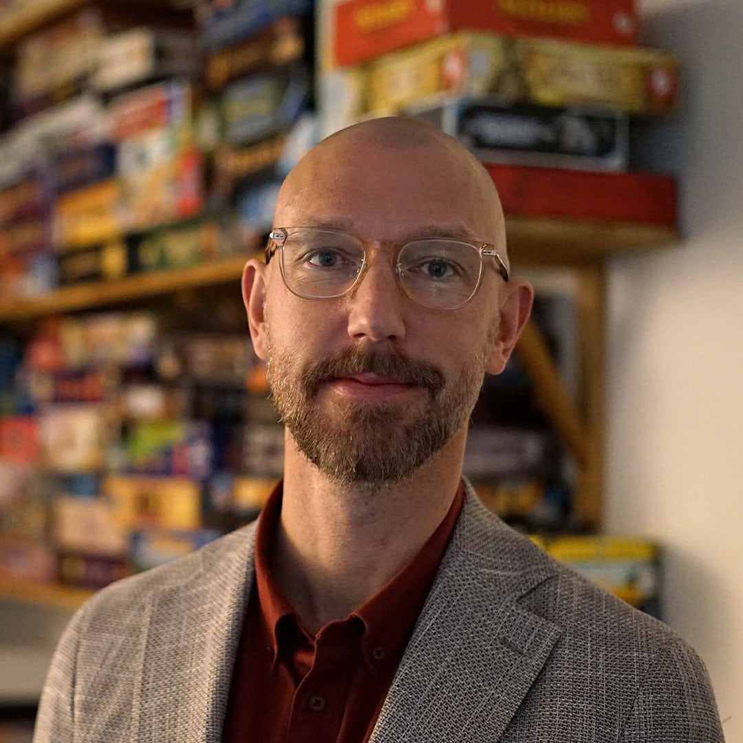 A man with a bald head, glasses, and a beard wearing a light-colored blazer and a red shirt stands in front of shelves filled with stacked board games.