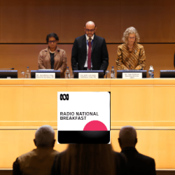 Four people stand behind a long panel table on a stage, facing an audience. Nameplates and microphones are in front of them. An overlay for “Radio National Breakfast” appears in the center foreground.