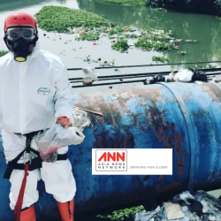 A person in a hazmat suit, helmet, gloves, and a respirator mask stands on a large blue pipe over a polluted river, holding plastic bags. Trash and debris are scattered around. ANN Asia News Network logo is visible in the foreground.
